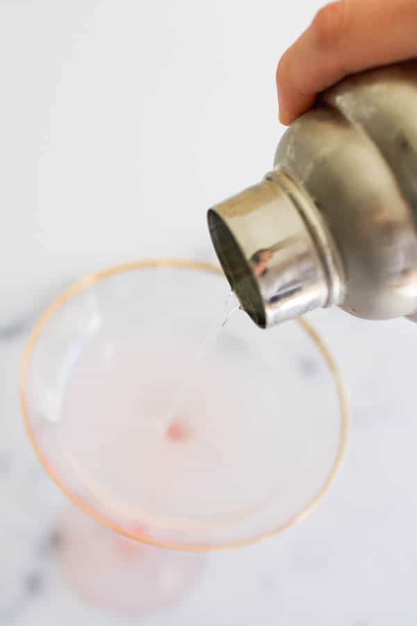 Pouring a vodka gimlet into a pink coupe glass from a cocktail shaker.