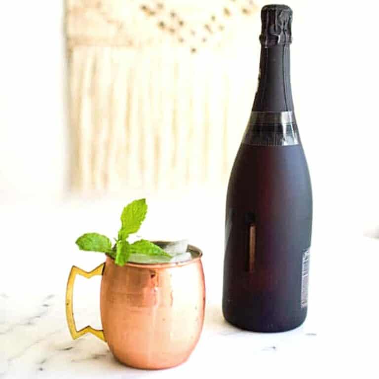 A bottle of champagne on a table next to a copper mug with a cocktail with a mint garnish.