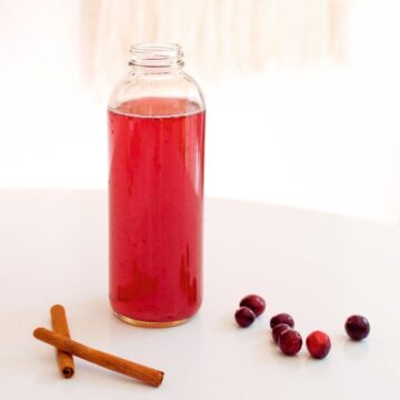 Spiced cranberry juice in a glass bottle next to cinnamon sticks and fresh cranberries.