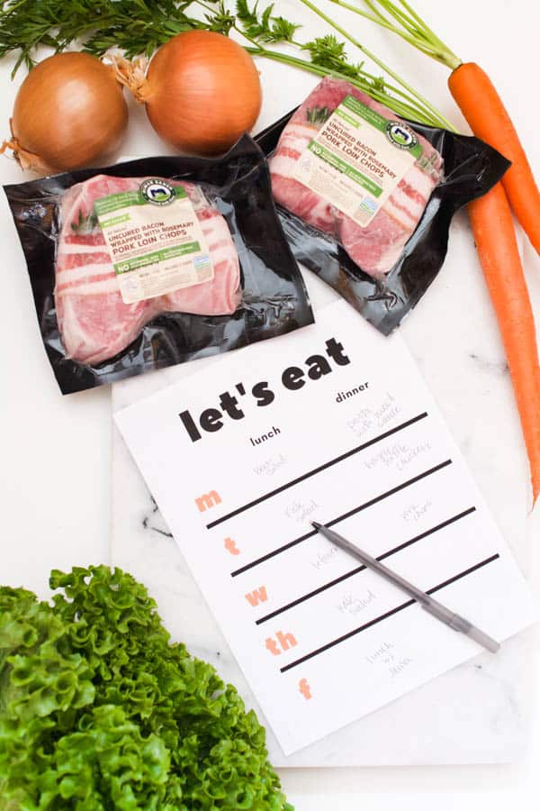 A meal planner on a cutting board next to packages of pork chops and carrots.
