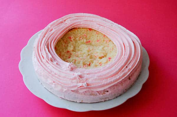 cake on a plate with pink frosting on part of the top with a hole in the middle to place candy before being topped by a chocolate dome