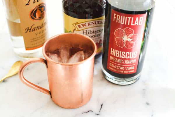 Copper Moscow Mule mug on a cutting board next to ingredients for a Hibiscus Moscow Mule.
