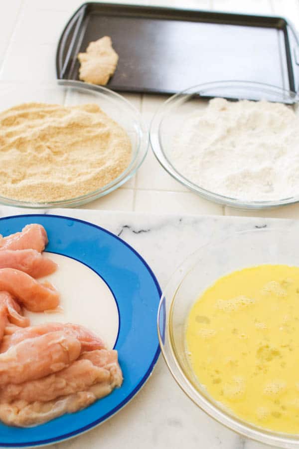 Dishes holding bread crumbs, flour and egg next to a plate of chicken tenders for making homemade breaded chicken tenders. 