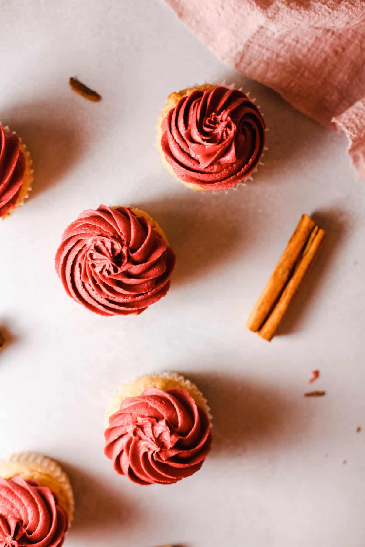Overhead view of cupcakes topped with red buttercream on a table next to cinnamon sticks.