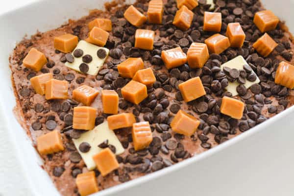 Baking dish holding chocolate cake mix, caramel candies, chocolate chips and pats of butter.