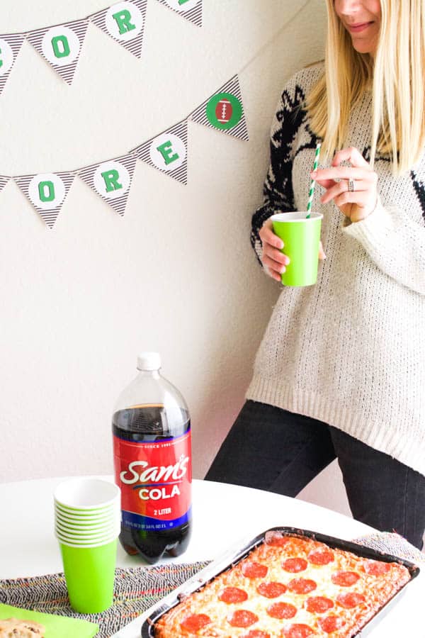 Woman holding a green cup with straw behind a table with a bottle of Sam\'s cola and a frozen pizza.