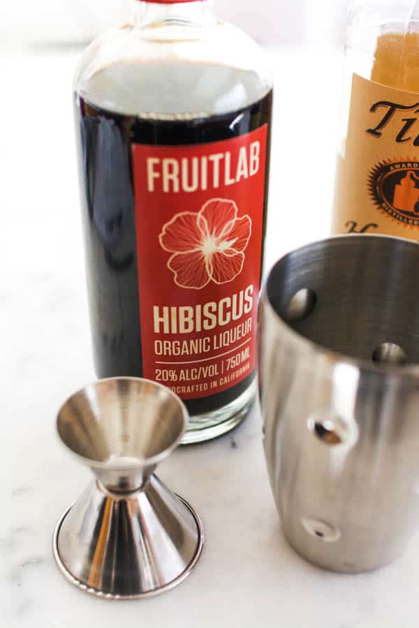 jigger and cocktail shaker on a counter next to ingredients to make a hibiscus martini