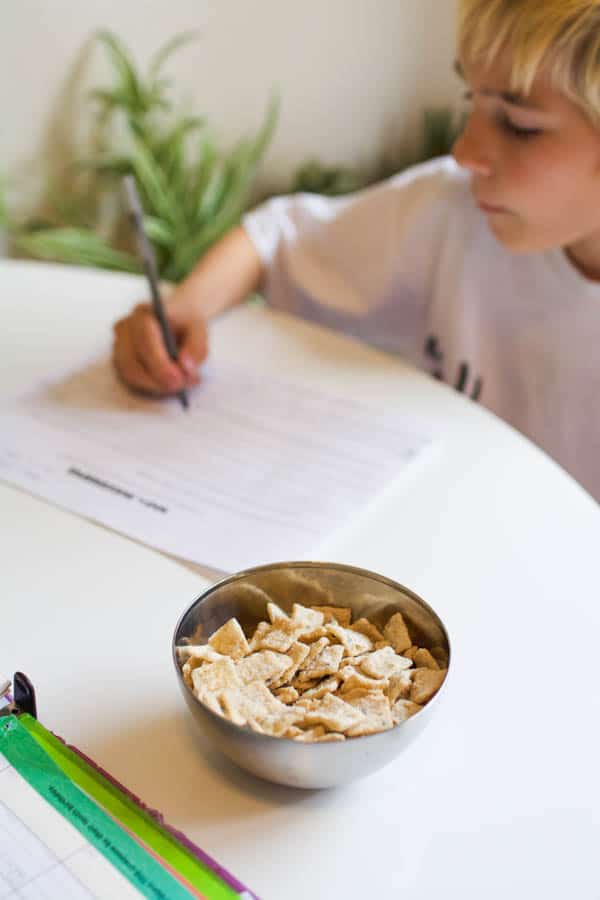 Boy sitting at table doing homework and eating cereal for a snack.