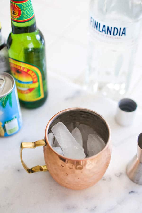 Copper mule mug with ice in it next to other ingredients on a cutting board.