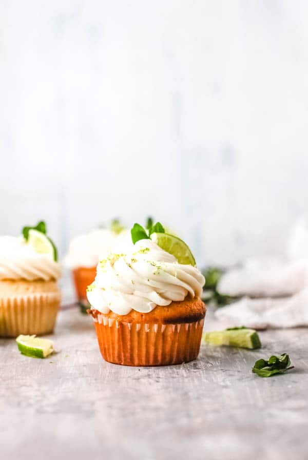 Side view of frosted cupcakes topped with lime zest, a lime wedge and some mint. 