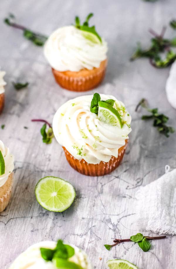 Frosted cupcakes on a table with limes slices and mint around.