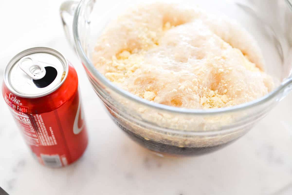 A can of Coke next to a bowl with cake mix and soda in it.
