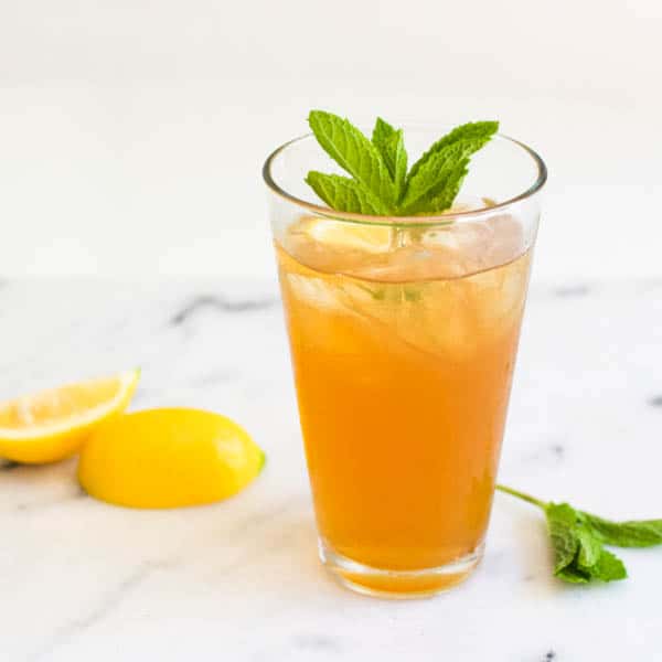 Tall glass of iced tea with mint garnish and lemon wedges next to it.