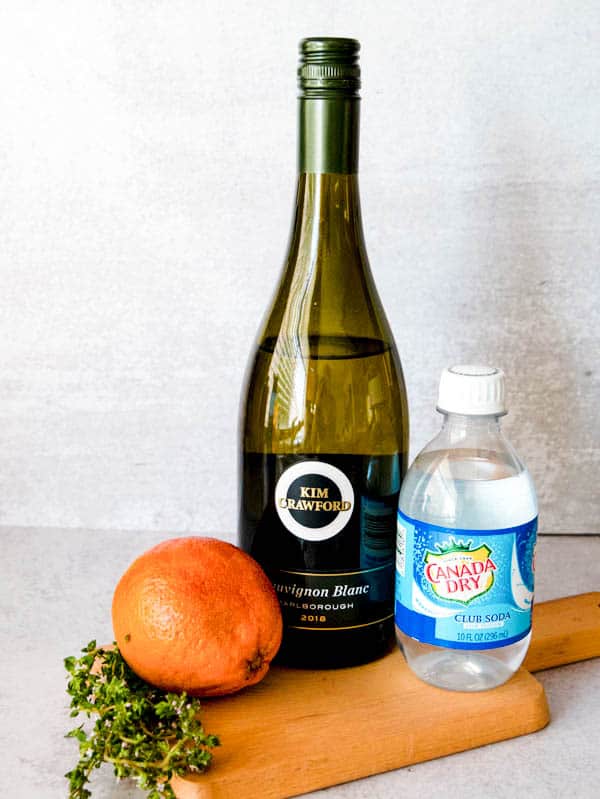 Ingredients for a white wine spritzer with thyme, a blood orange a bottle of sauvignon blanc and club soda.