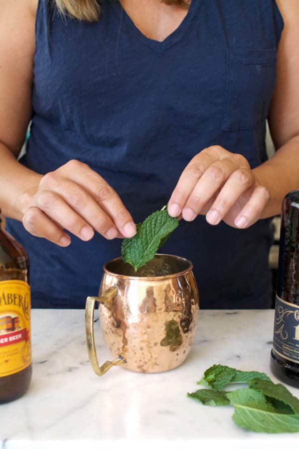 Woman adding a fresh mint leaf to a copper mug for a Rum Mule cocktail.