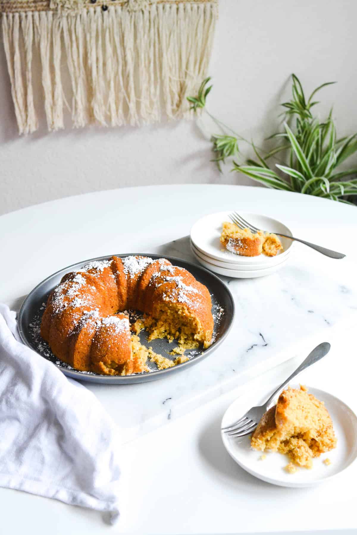 A bundt cake topped with powdered sugar on a table next to dessert plates with cake on them.