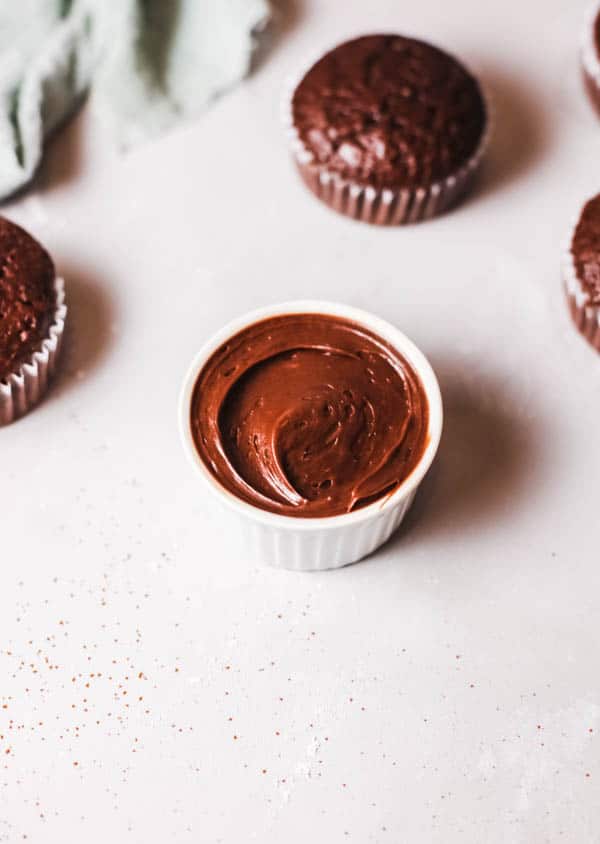 Chocolate frosting in a bowl with a few chocolate cupcakes around it. 