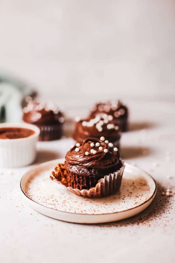 Frosted chocolate cupcake with white round sprinkles on a plate with the wrapper partially removed.