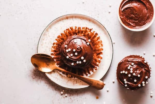 Frosted chocolate cupcake on a plate with a spoon and round white sprinkles. 