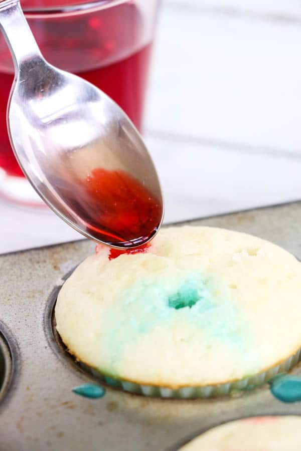 a spoon adding liquid red Jello to a poked hole in the top of a baked cupcake