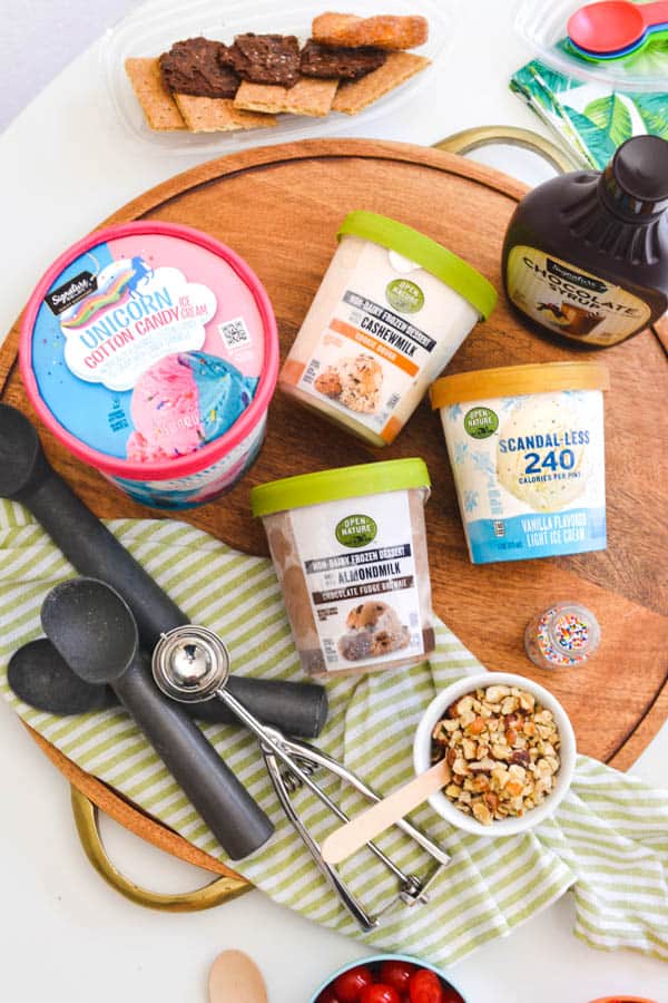 A variety of ice cream laying on a cutting board next to a bottle of chocolate sauce and bowl of nuts.