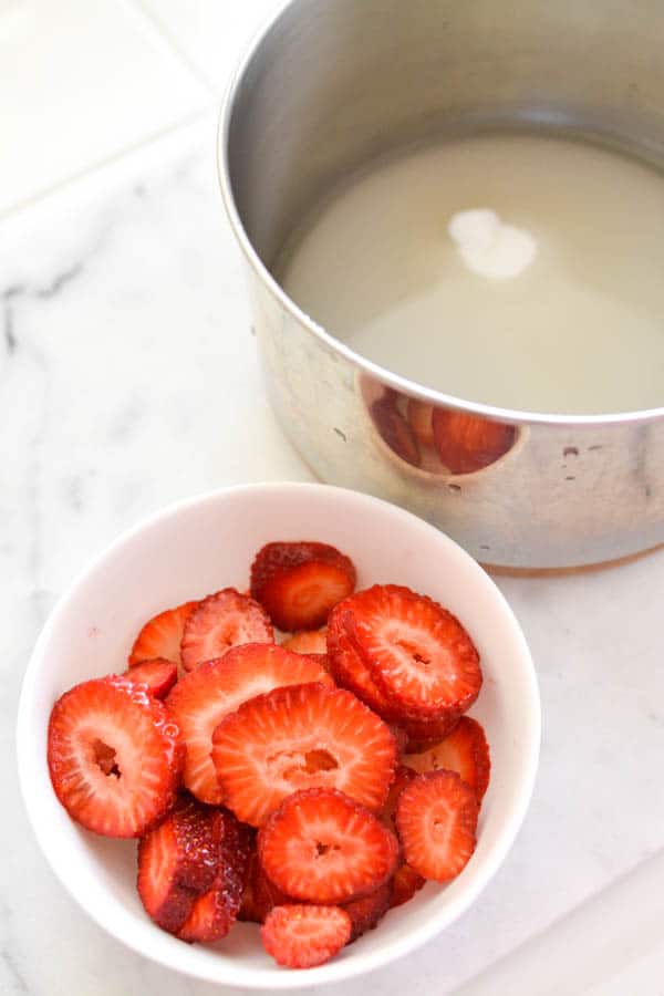 Sauce pan with sugar and water next to a bowl of sliced fresh strawberries.