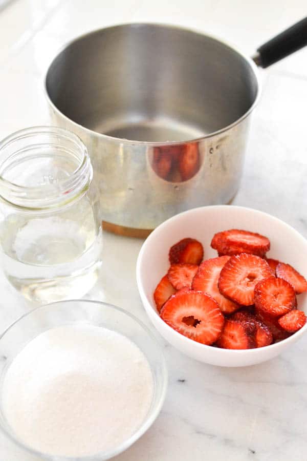 Sauce pan next to a bowl of fresh strawberries, sugar and water.