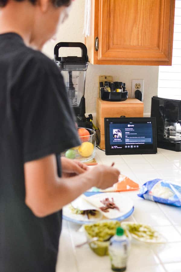 Kid making lunch at the kitchen counter with a Portal device behind him. 