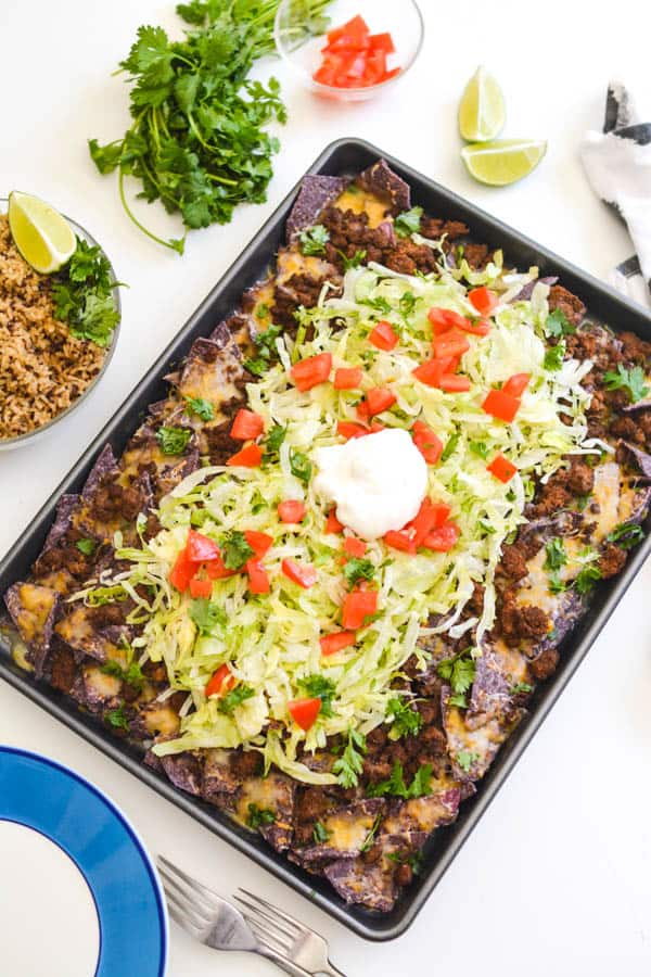 Nachos on a sheet pan with shredded lettuce and sour cream on a table.
