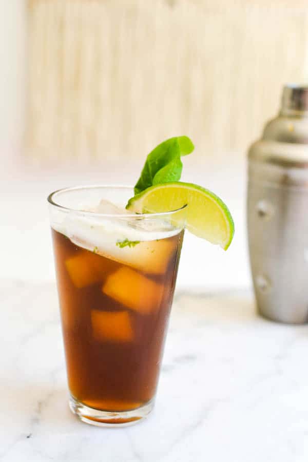 A glass of iced tea on a table with a cocktail shaker behind it and garnished with a lime wedge and fresh lime.