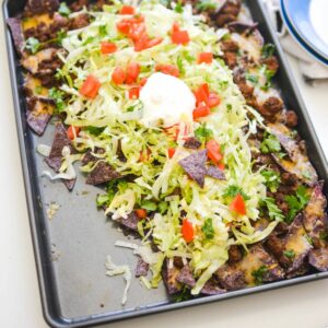 Close up of a sheet pan full of nachos topped with shredded lettuce, sour cream and tomatoes.