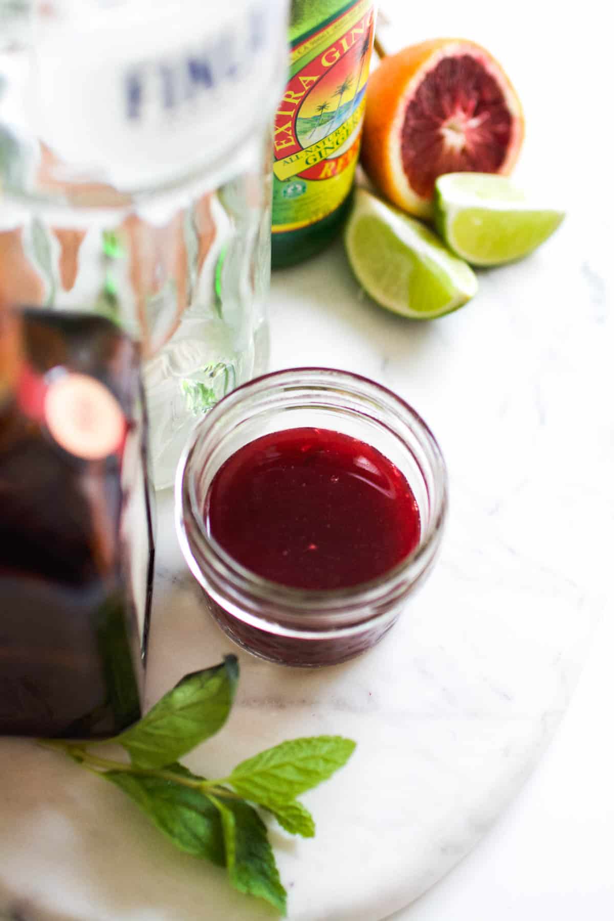 Close up of fresh squeezed blood orange juice in a small bowl next to fresh fruit and a bottle of vodka.