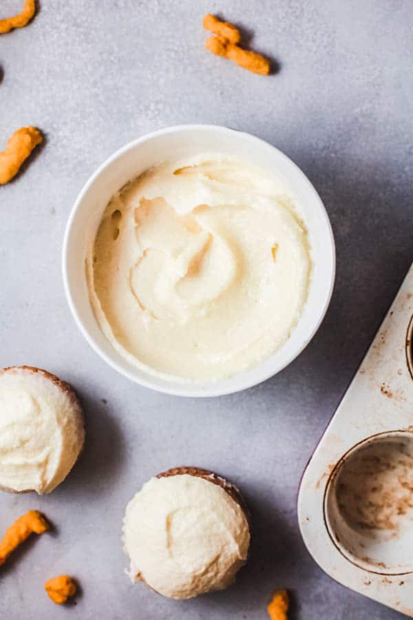 A bowl of cream cheese frosting on a table next to cupcakes.