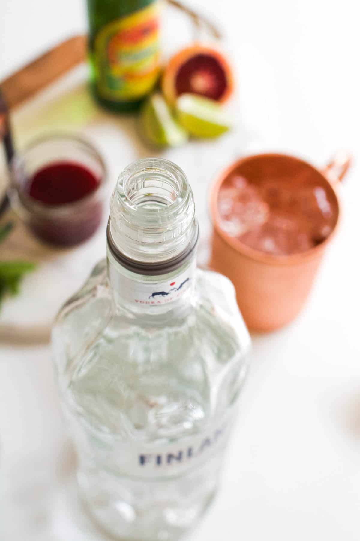 Close up of an open vodka bottle next to other ingredients for an orange mule.