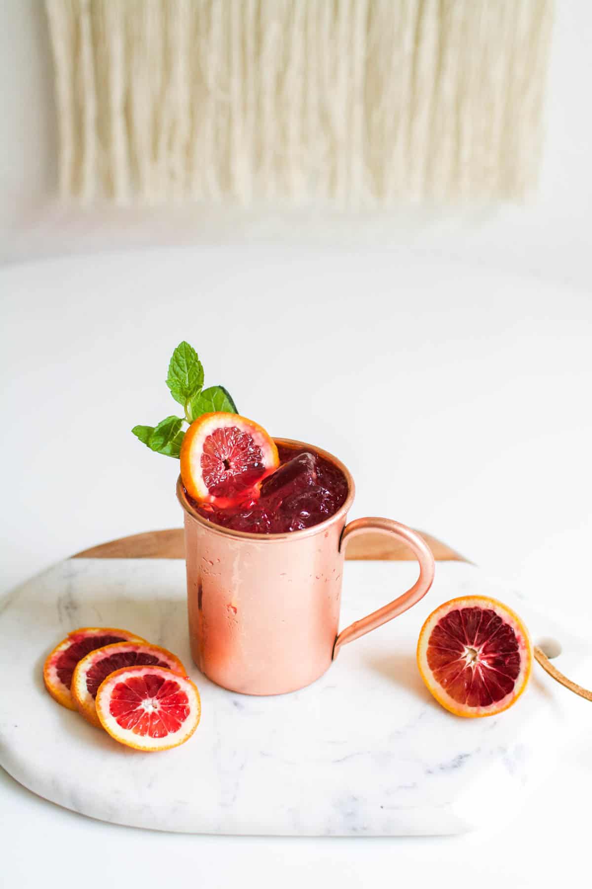A copper mug on a small marble tray with fresh blood oranges as garnish and around the glass on a table.