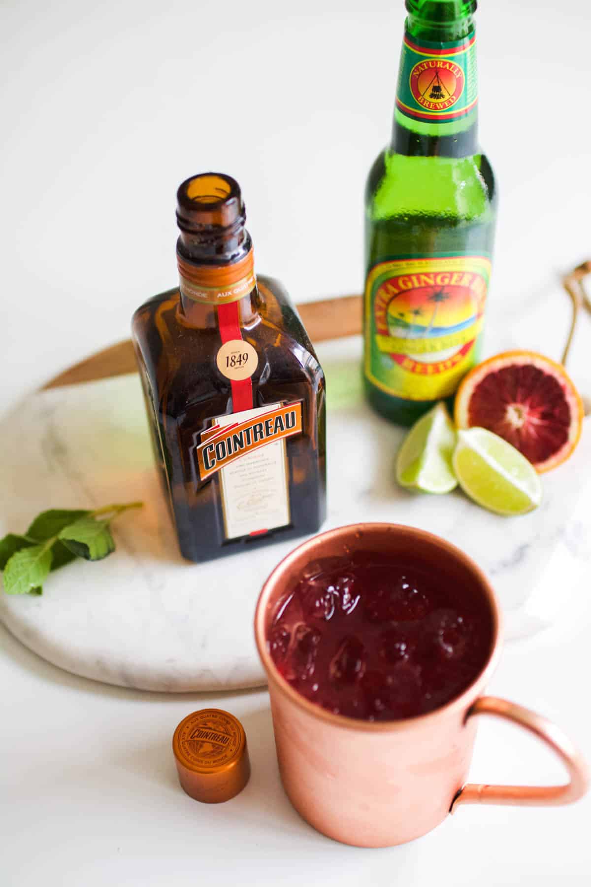 A copper mug on a table next to ginger beer, fresh fruit and Cointreau.