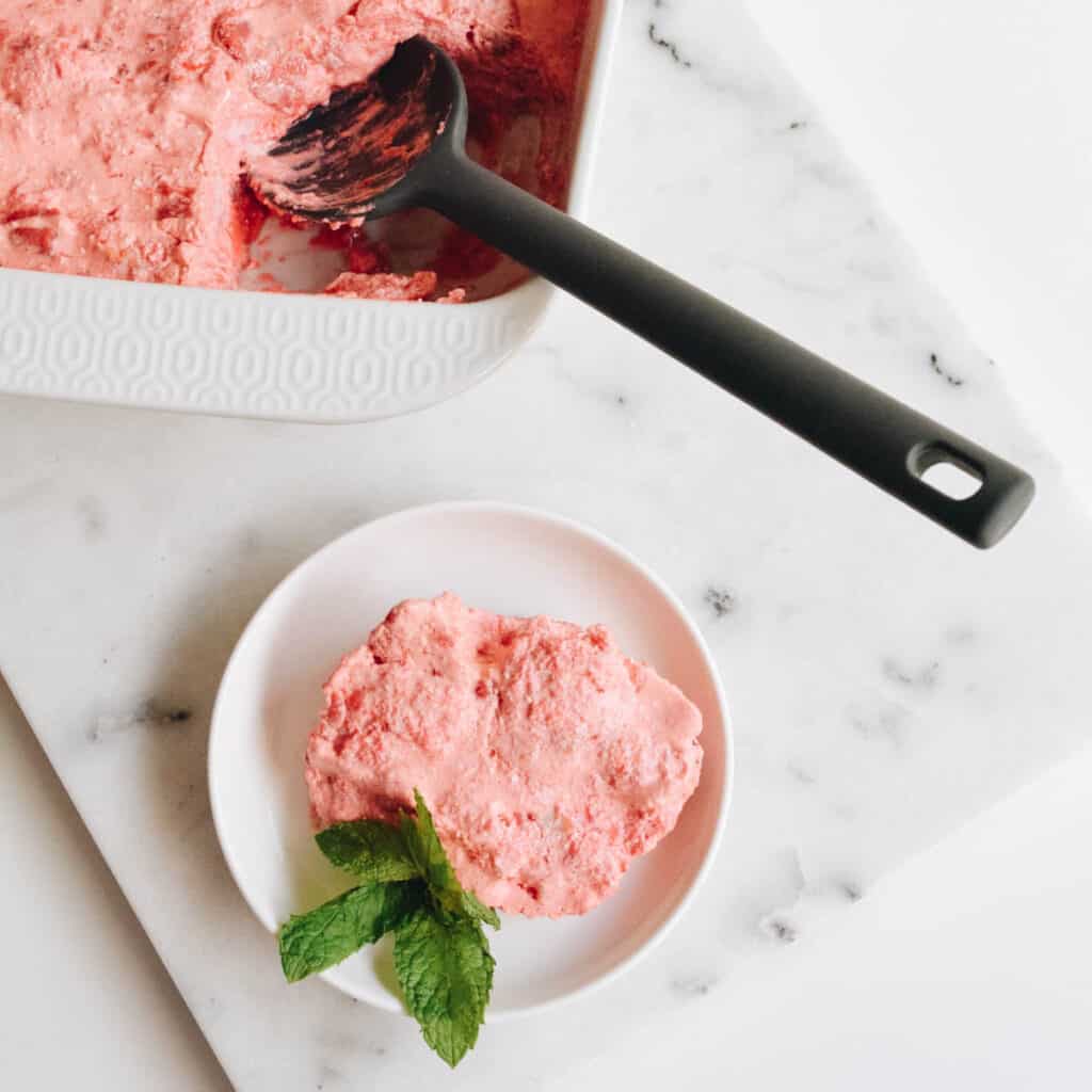 White baking dish with Strawberry Jello dessert with a small plate holding a serving in front of it on a table.