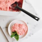 White baking dish with Strawberry Jello dessert with a small plate holding a serving in front of it on a table.