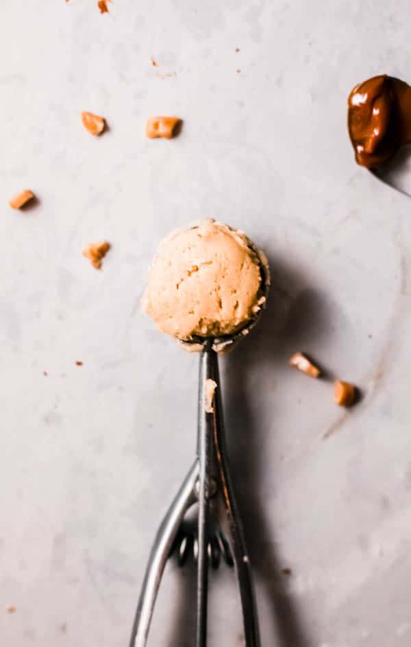 Small ice cream scooper tool with cookie batter on a table with butterscotch chips laying next to it.