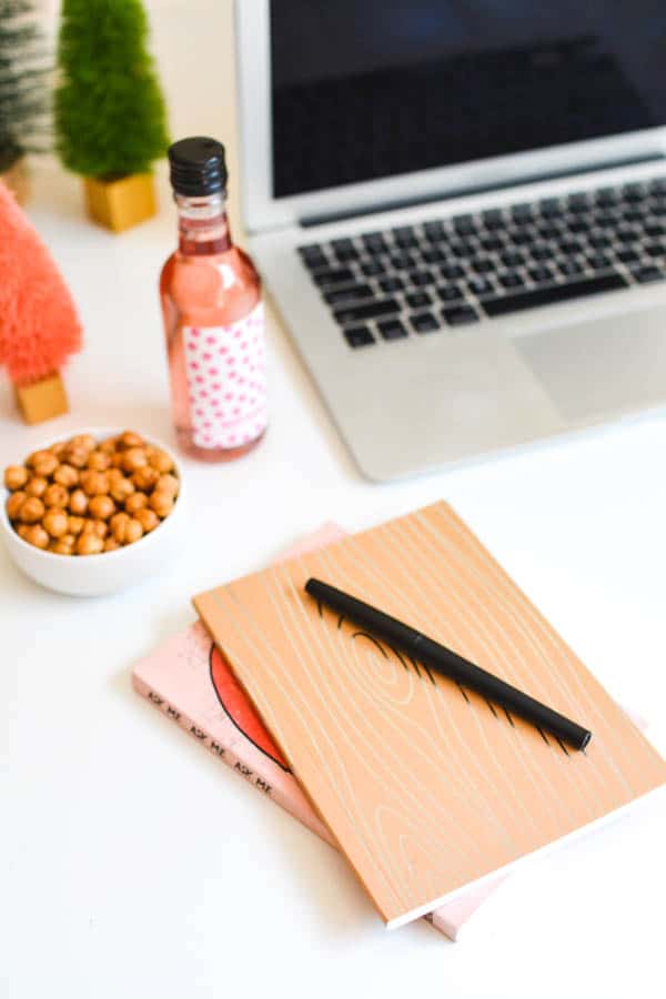 A book and notebook sitting on a table next to a laptop, a bowl with snacks and a mini bottle of wine.