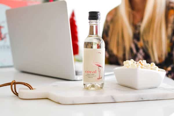 Close up on a mini bottle of white wine next to a bowl of popcorn with a girl on a laptop in the background.
