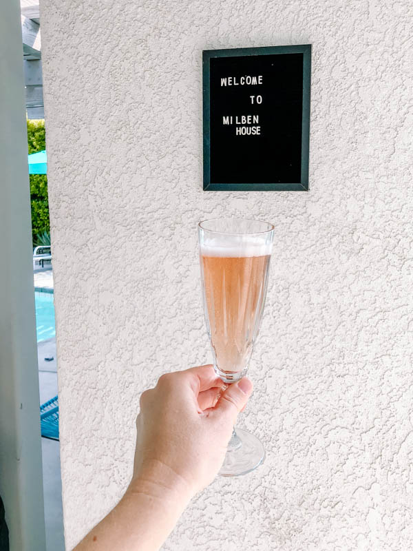 Girl\'s hand holding a glass of champange in front of a sign that says \"Welcome to Milben House.\" 