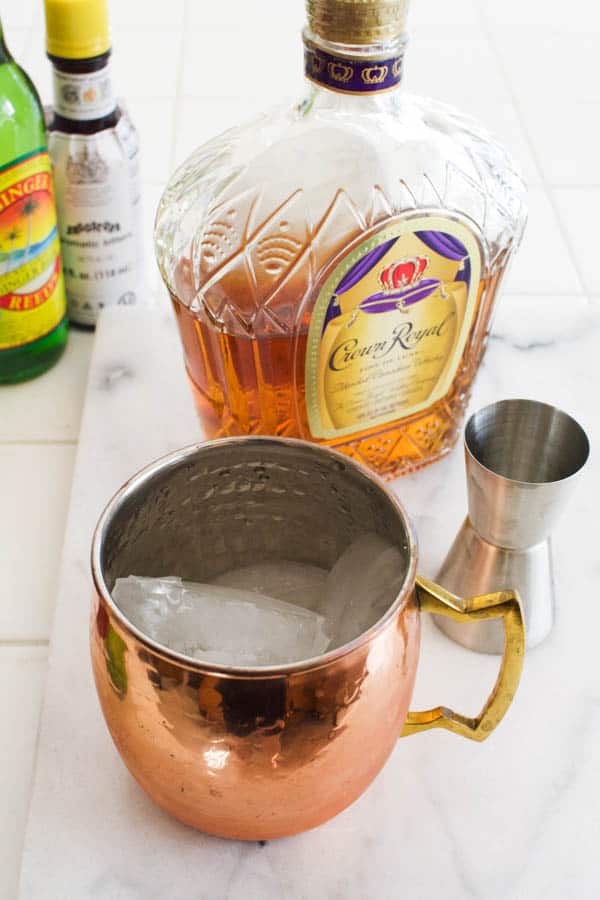Copper Moscow Mule mug on a cutting board with ice next to whiskey and a jigger.