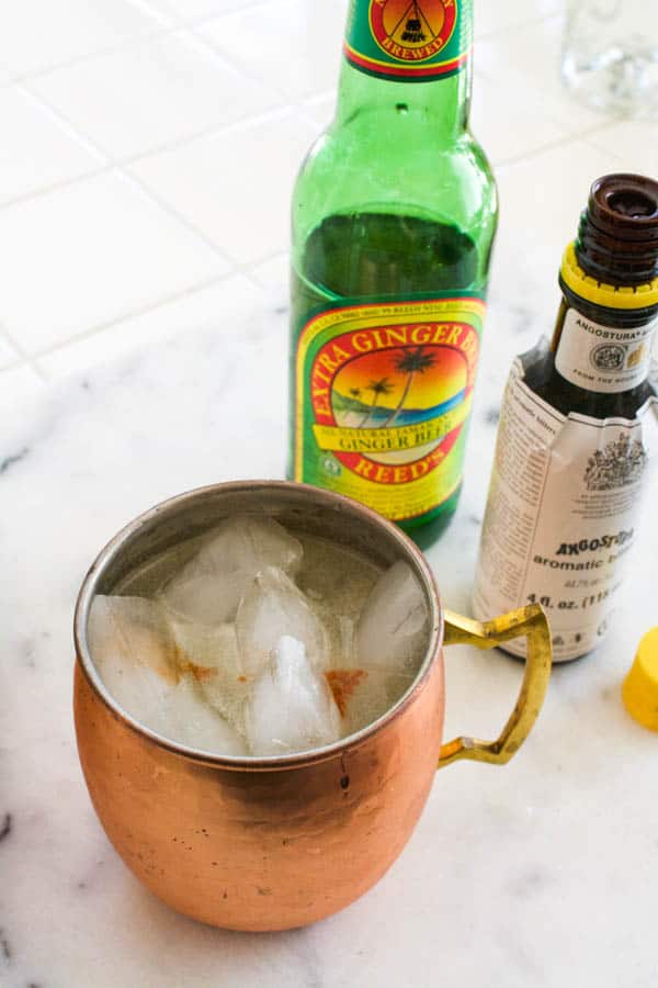 A copper mug next to ginger beer and bitters on a cutting board.