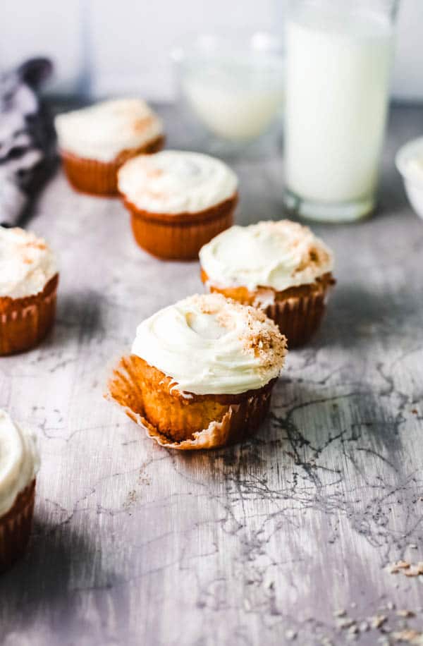 Frosted cupcakes on a table with a glass of milk in the background.