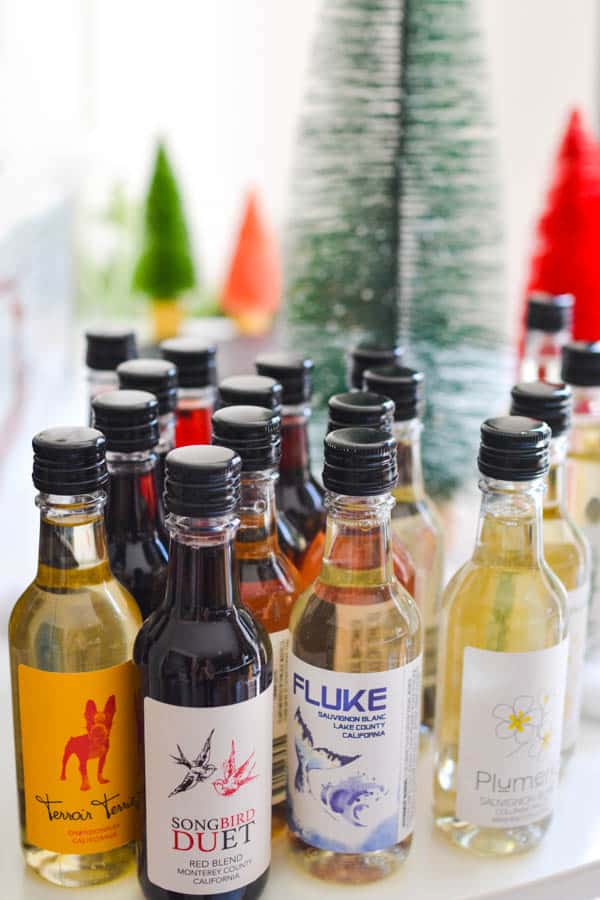 Mini bottles of wine, with decorative labels, grouped on a table with bottle brush holiday trees in the background. 