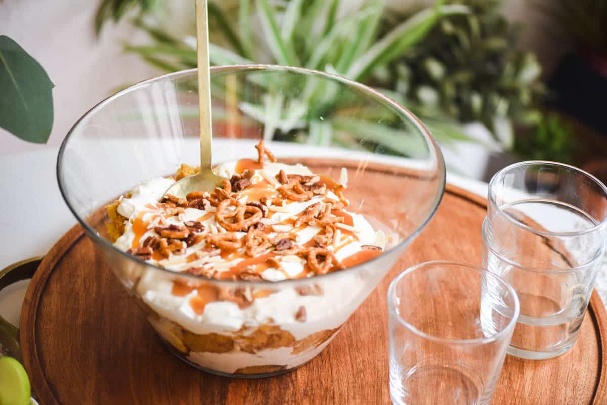 Close up of a dessert topped with nuts, pretzels and caramel with serving glasses next to it.