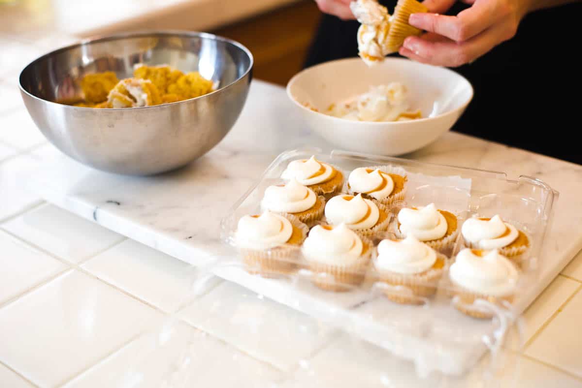 Tray of mini cupcakes on a cutting board with a woman scraping the frosting off of some in the background.