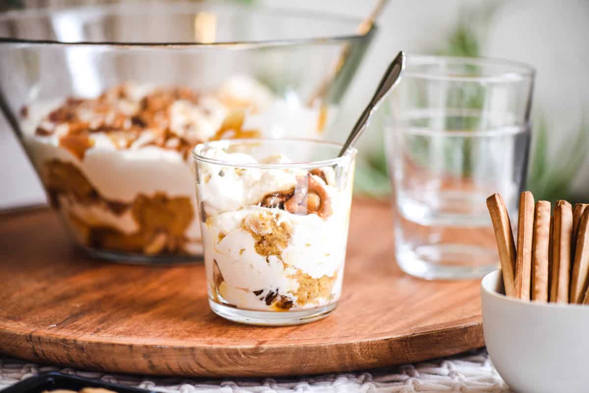 Close up of an individual glass holding a serving of pumpkin trifle on a tray with a bowl of the dessert in the background.