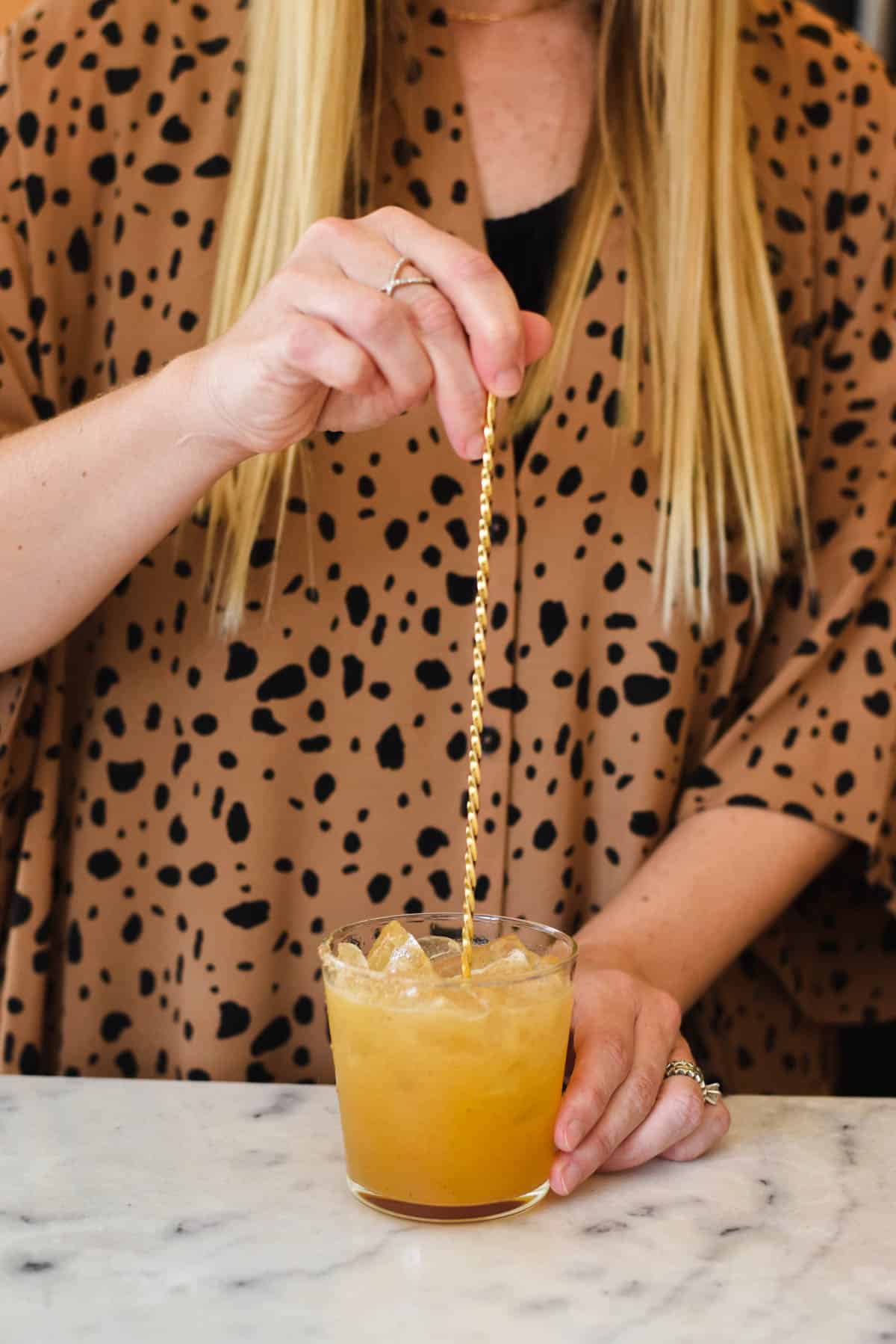 Woman using a bar spoon to mix a cocktail on a counter.
